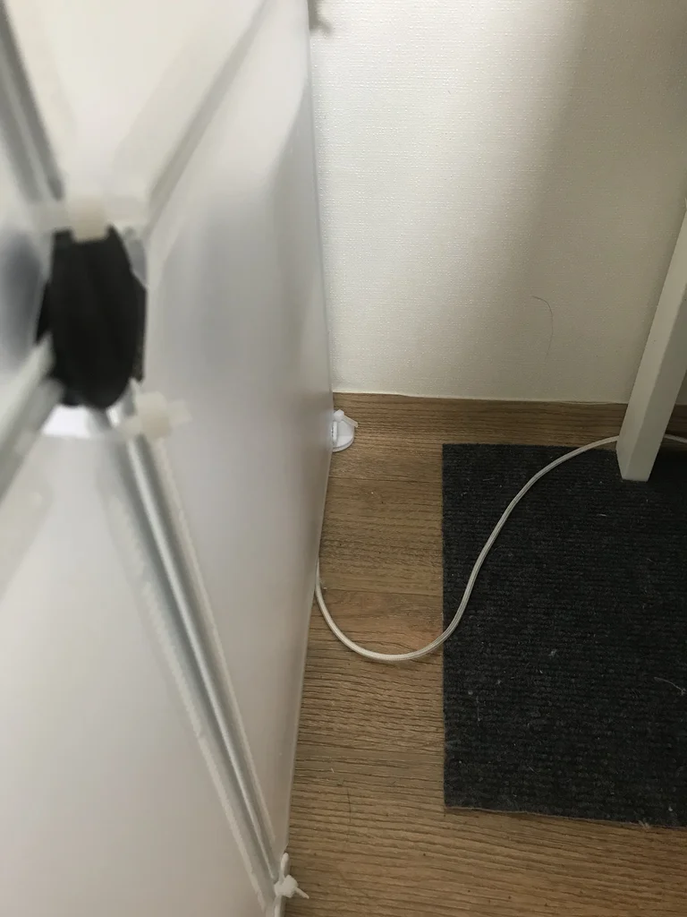 The feeder's cable running outside the opaque panel wall of a DIY rabbit enclosure, along the wall at floor level